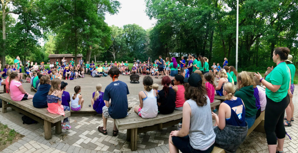 A panoramic photo shows a large group of kids and counselors gathered around a plaza at camp, sitting on wooden benches.