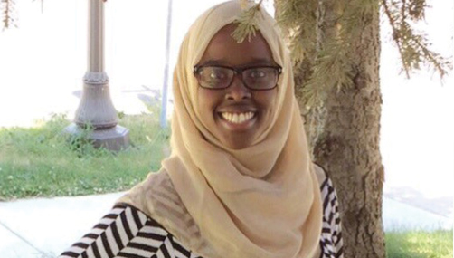 A young woman wearing a hijab and glasses smiled at the camera by a tree