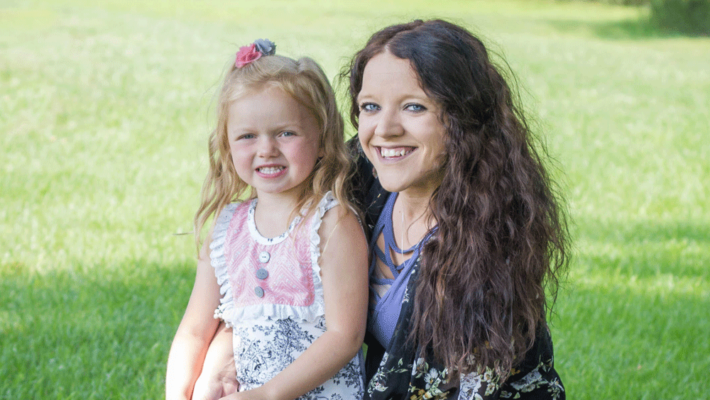 A woman with long curly hair and a young girl on her lap both smile at the camera.