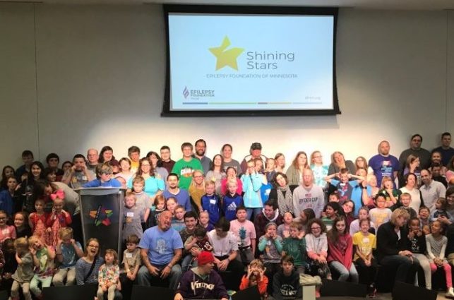 A large group of children with epilepsy and their families, in front of a screen showing the Shining Stars logo.