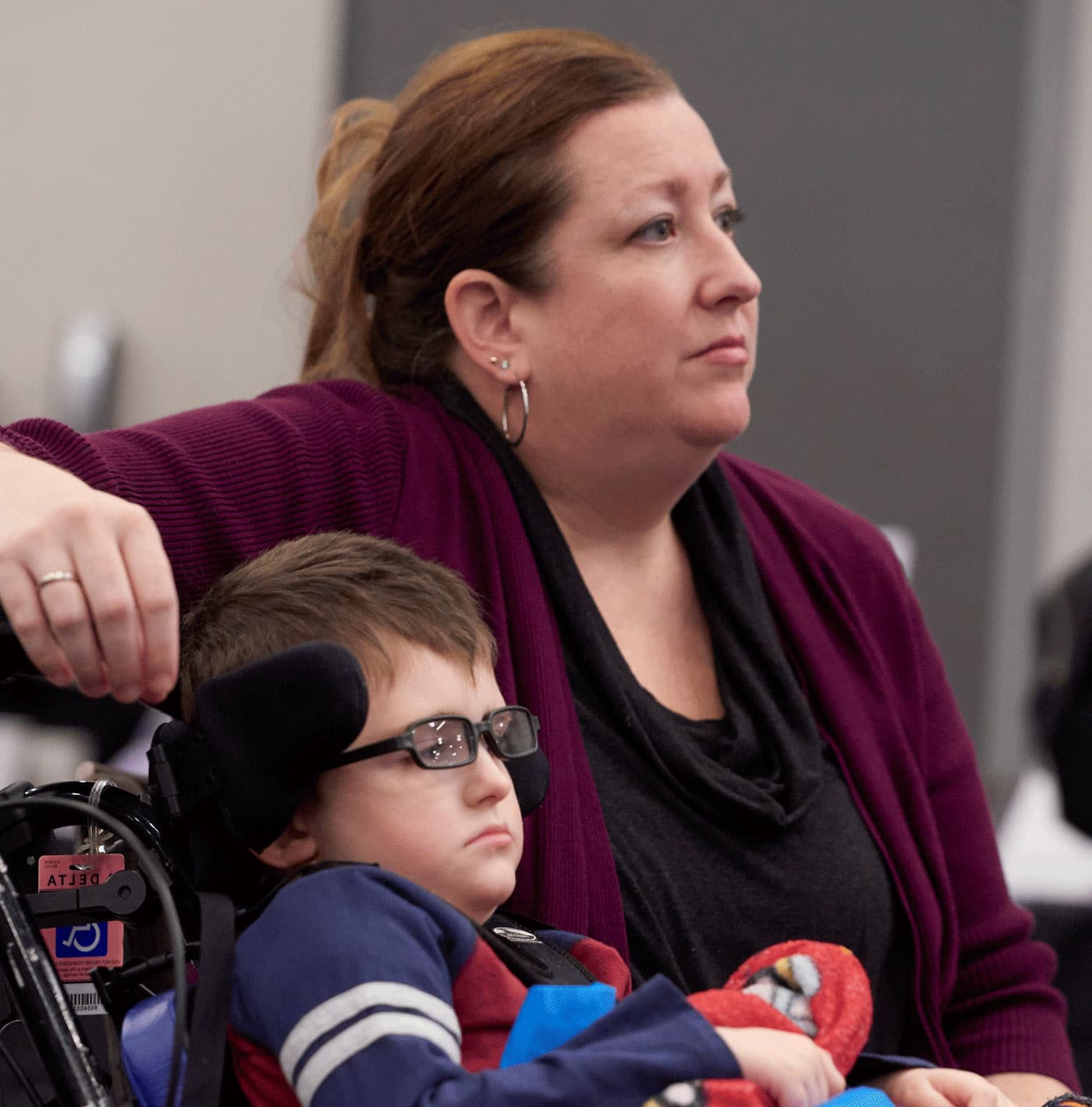 A woman in a maroon sweater leans an arm over her young son's wheelchair, listening intently to someone off camera.