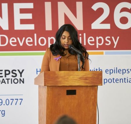 Woman in a brown blazer and purple ribbon gives a speech at a podium in front of a "one in 26" banner.