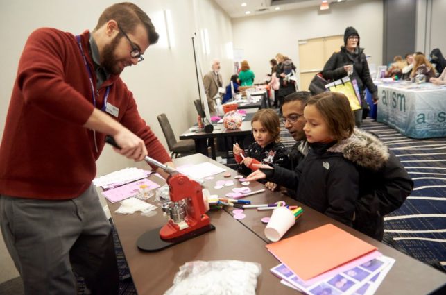 Man in a red sweater uses a red machine to make a button while three children look on from the other side of the table.