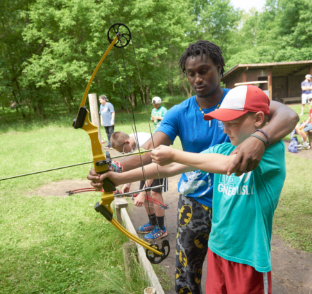 A camp counselor helps a younger boy draw his bow on an archery feild