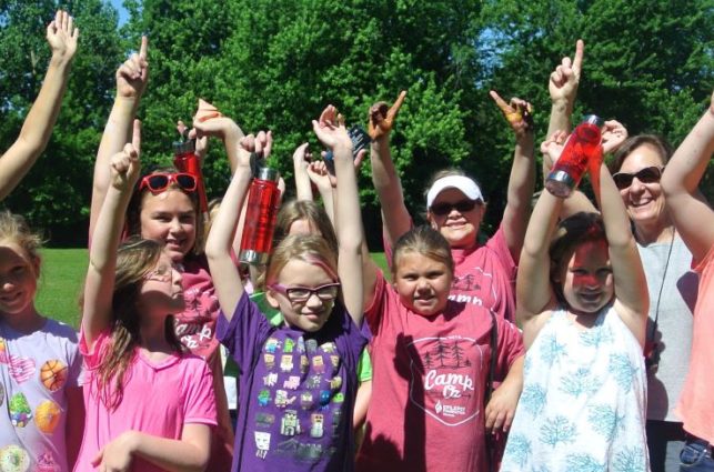 A group of kids in brightly colored tshirts stand together hold up their index fingers to indicate "one."