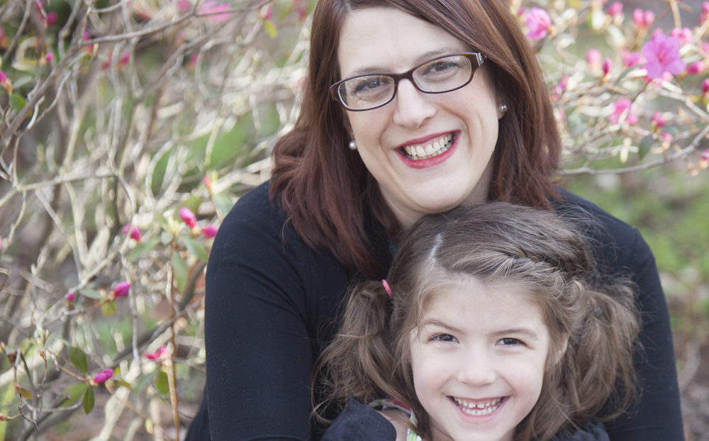 A mother hugs her daughter in front of a rosebush.