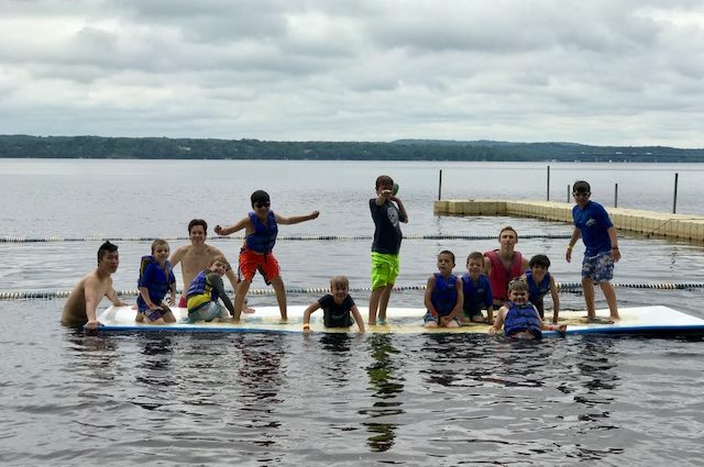 A group of kids poses and goofs around on a raft in a lake at camp.