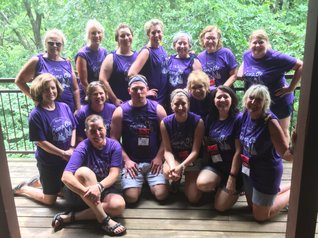 A group of 20 camp counselors and nurses group together on a balcony wearing matching camp oz t-shirts. 