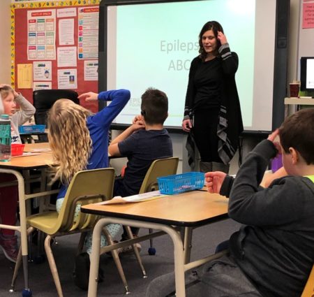 A woman stands at a projector and talks to students in a classroom.