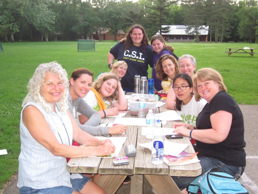 A group of people lean in over a picnic table in the sun at Camp Oz.