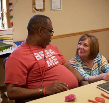 A woman crouches next to a man sitting at a table, and they smile and talk.