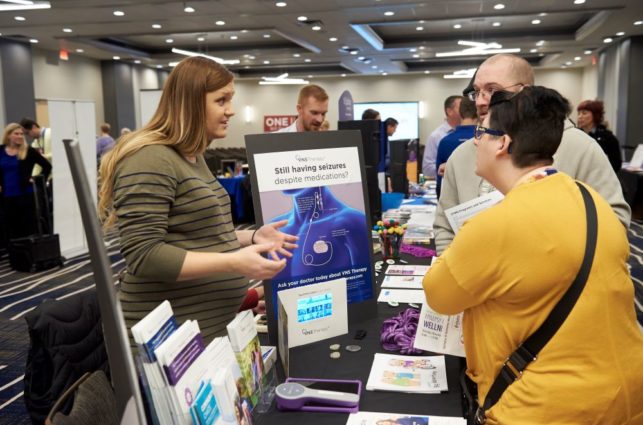 A woman at a booth explains information about epilepsy to two older people.