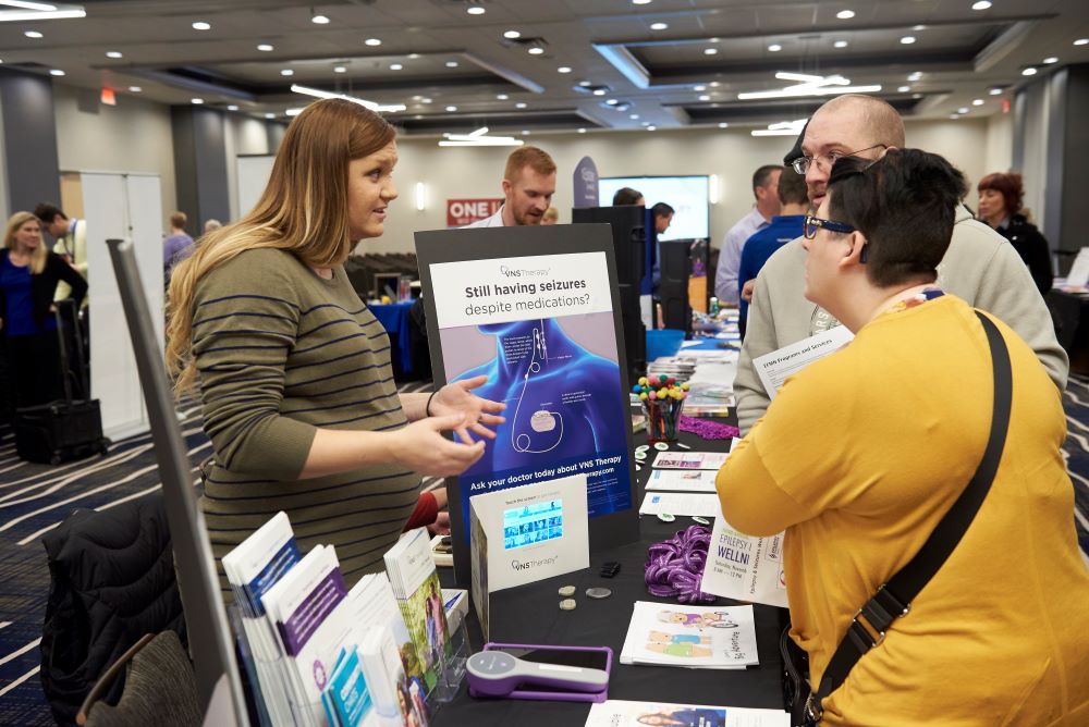 A woman at a booth explains information about epilepsy to two older people.