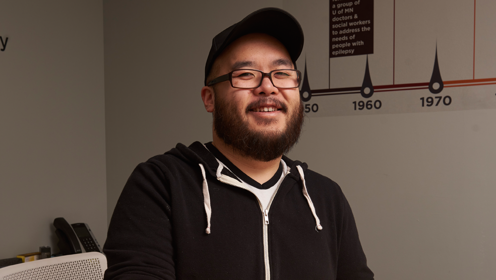 A man in a hoodie and baseball camp sits in an office and smiles at the camera