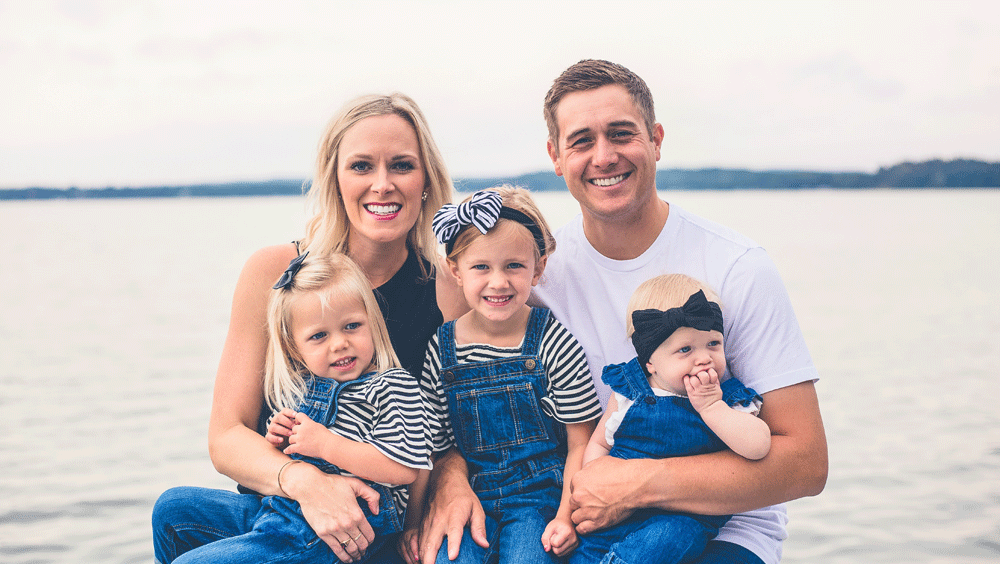 A woman and man with three young children wearing overalls pose for a portrait in front of a lake.