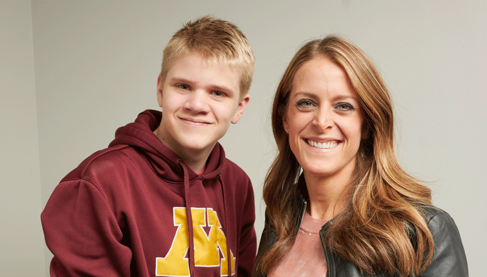 A young man with blond hair in a UofM sweater stands next to his mom, smiling. The family resemblance is apparent.