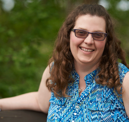 A woman with curly brown hair and sunglasses leans against a fence.