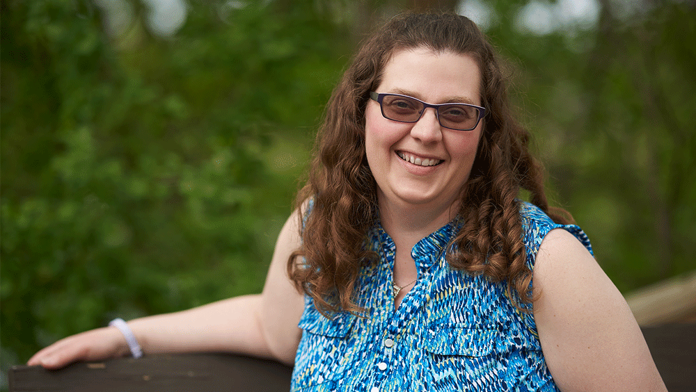 A woman with curly brown hair and sunglasses leans against a fence.