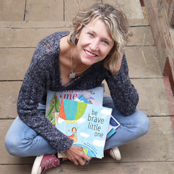 A woman with wavy blond hair sits crosslegged on the ground outside, holding a stack of children's books