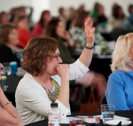 A woman sits in a crowd of people at a training and raises her hand to speak.