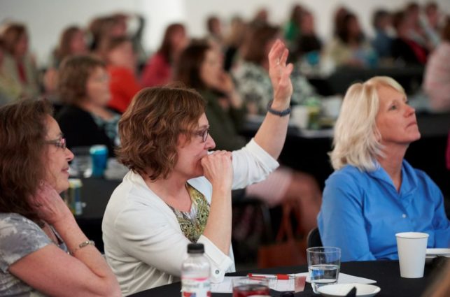 A woman sits in a crowd of people at a training and raises her hand to speak.