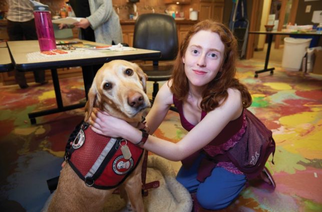 A young woman with red hair smiles at the camera while hugging her seizure dog, a yellow lab wearing a red "Can Do Canine" working vest