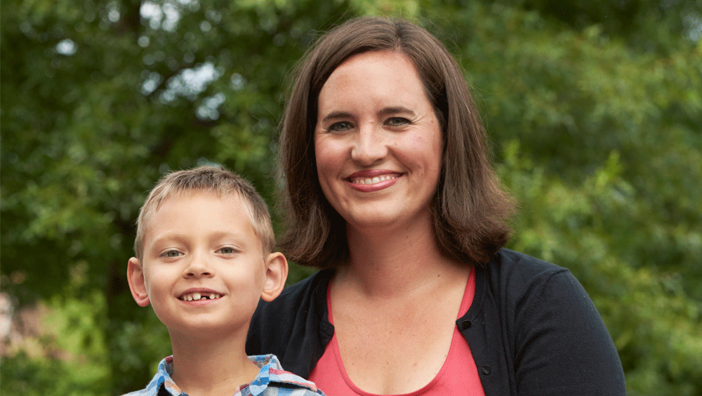 A small boy sits with his mom in a park, smiling at the camera to reveal a missing front tooth