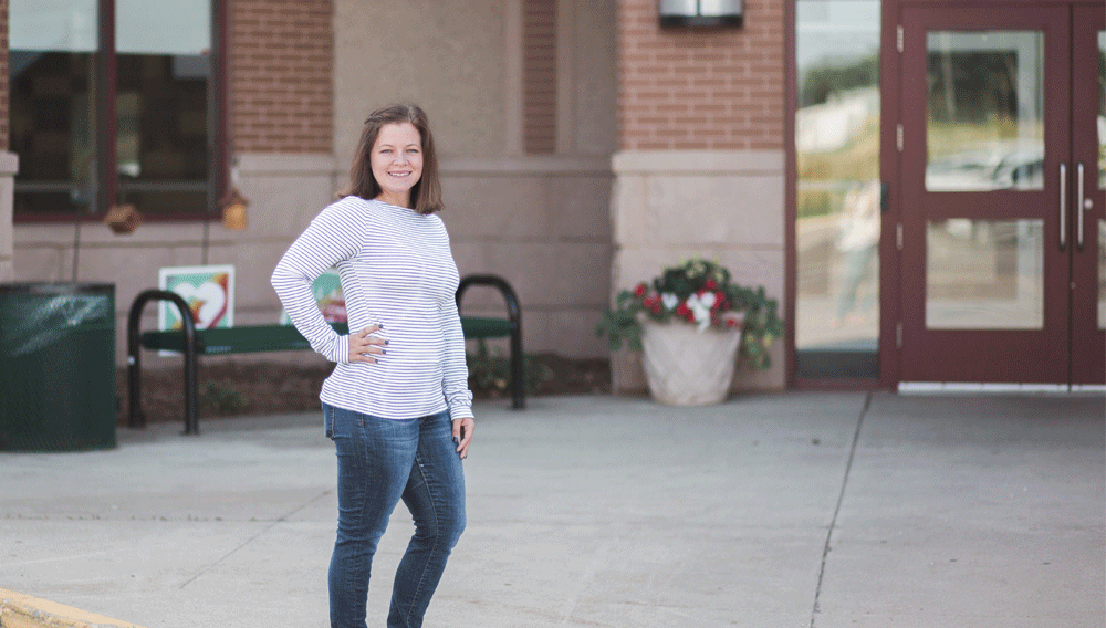 A woman in a striped shirt stands with her hand on her hip in front of a school building.