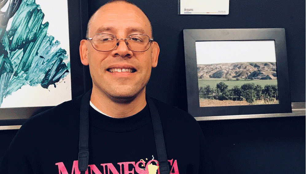 A man in a Minnesota sweatshirt holds a camera and stands next to one of his photographs.