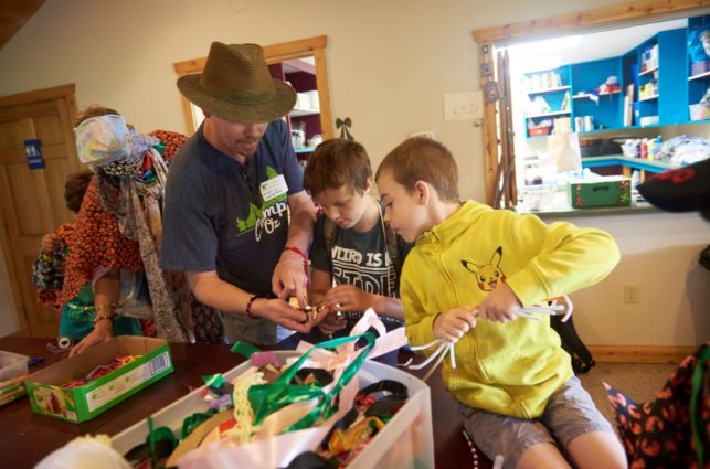 An adult assists two children with crafts in a classroom at camp.