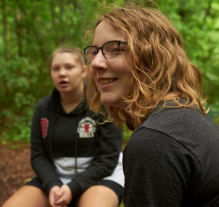 Two young people sit in a wooded outdoor area, talking as part of a group.