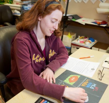 A young woman works on an art project at a table with papers and art supplies.