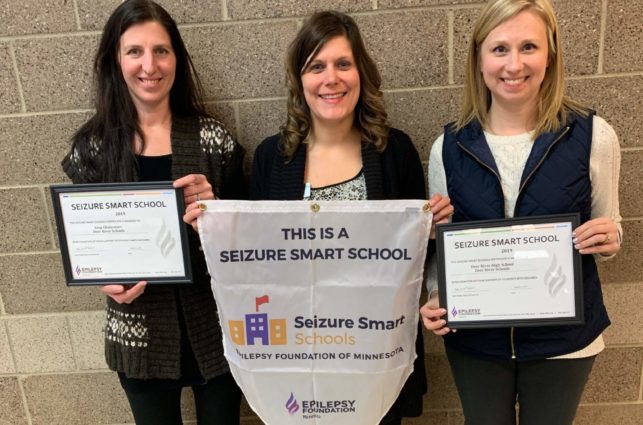 Three women stand against a wall, displaying certificates and banners showing their school is a Seizure Smart School.