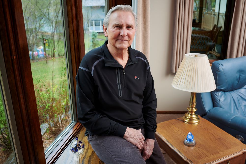 An older man in a black zip-up sits in front of a picture window in his living room