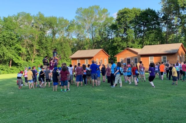 A large group of campers gather in a field in front of cabins at Camp Oz