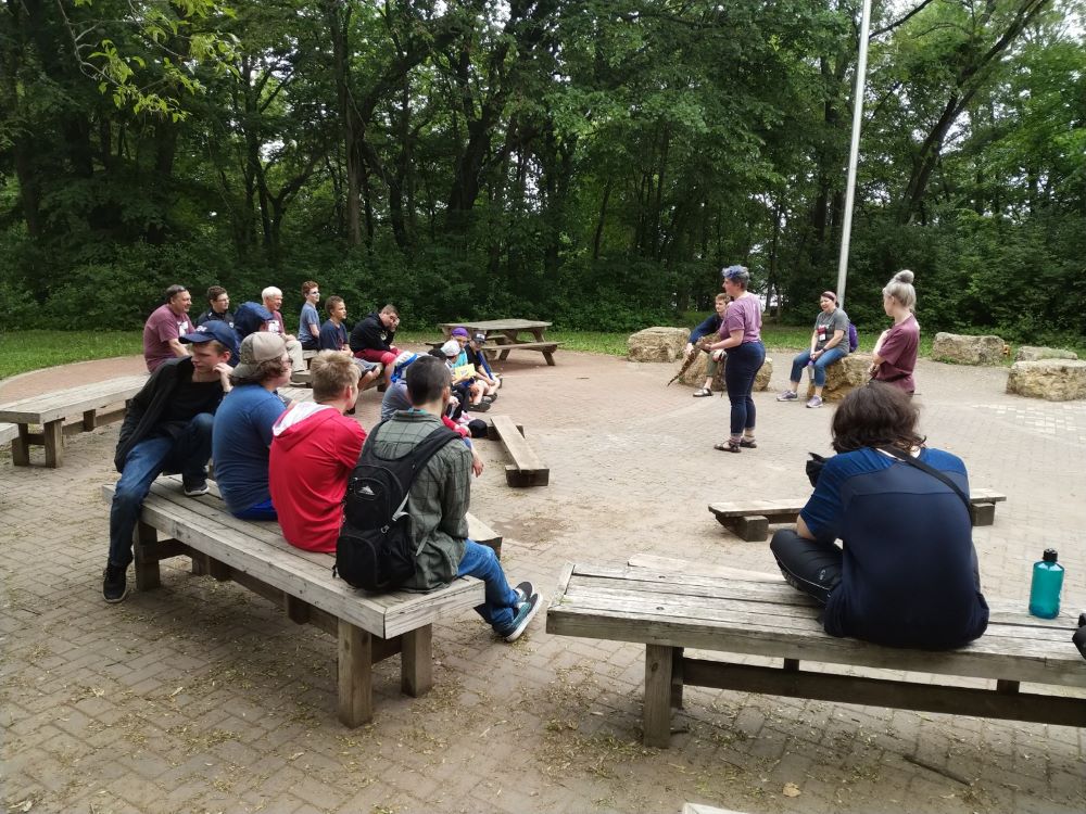 A group of older campers sit on benches around a campfire