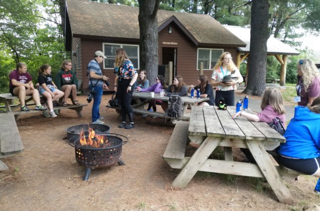 Campers sit on a ring of picnic tables around a campfire, socializing.