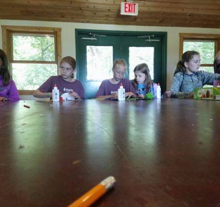A group of campers in matching camp shirts sit around a table working on crafts