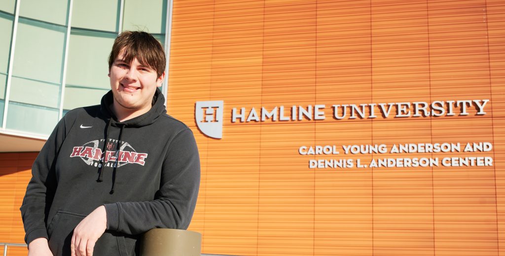 White male college student smiling in front of Hamline University sign