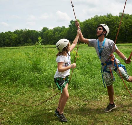 An adult and a child, both wearing helmets, work on a ropes course at camp.