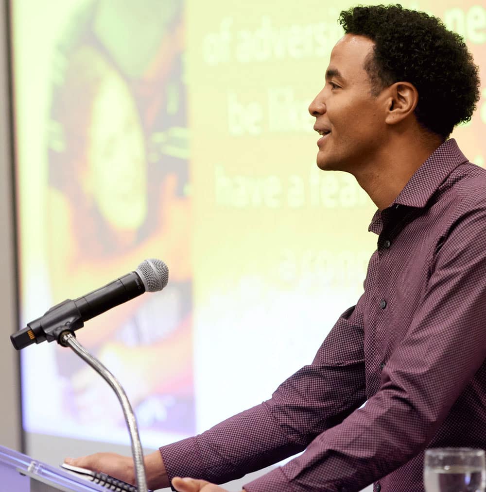 A man gives a speech at podium in front of a projector screen, smiling.