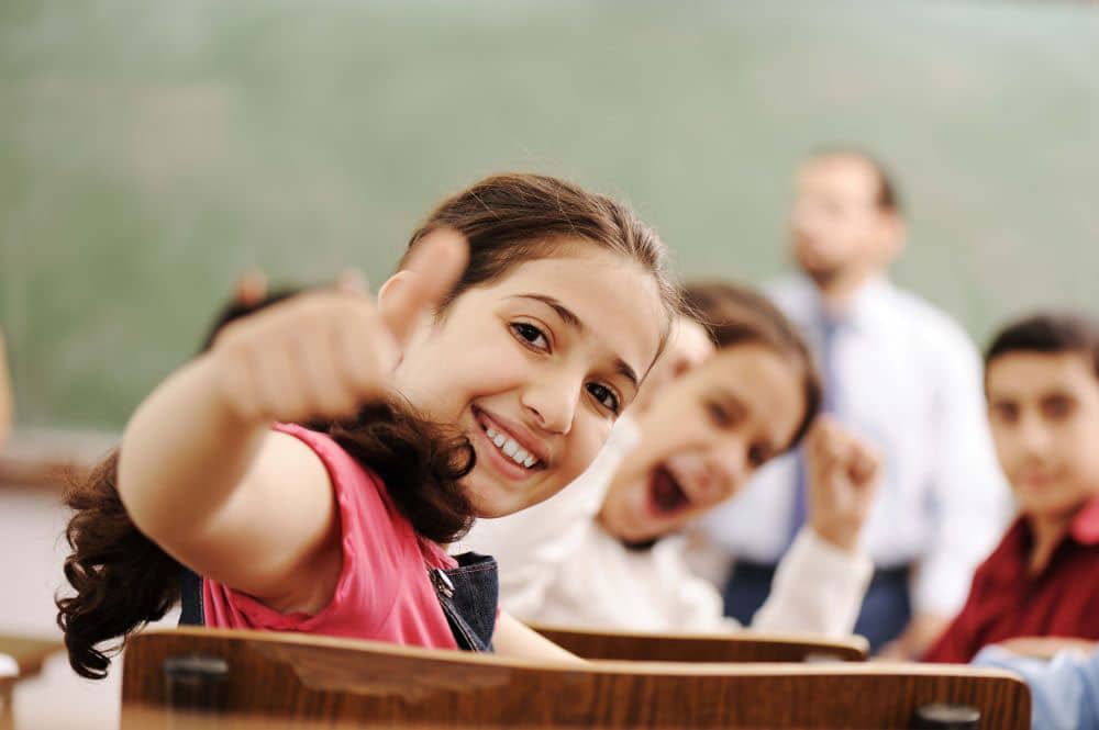 A girl gives a big smile and thumbs up to the camera. Behind her, more students smile and cheer, and a teacher stands at the front of the classroom.