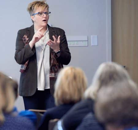 A woman with short blond hair and a black blazer counts off items on her fingers while giving a talk in front of a crowd of adults.