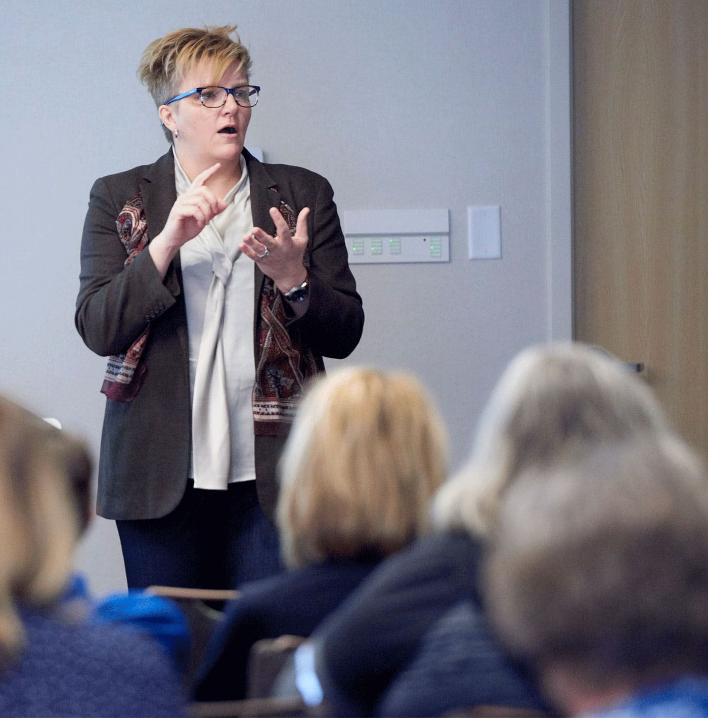 A woman with short blond hair and a black blazer counts off items on her fingers while giving a talk in front of a crowd of adults.