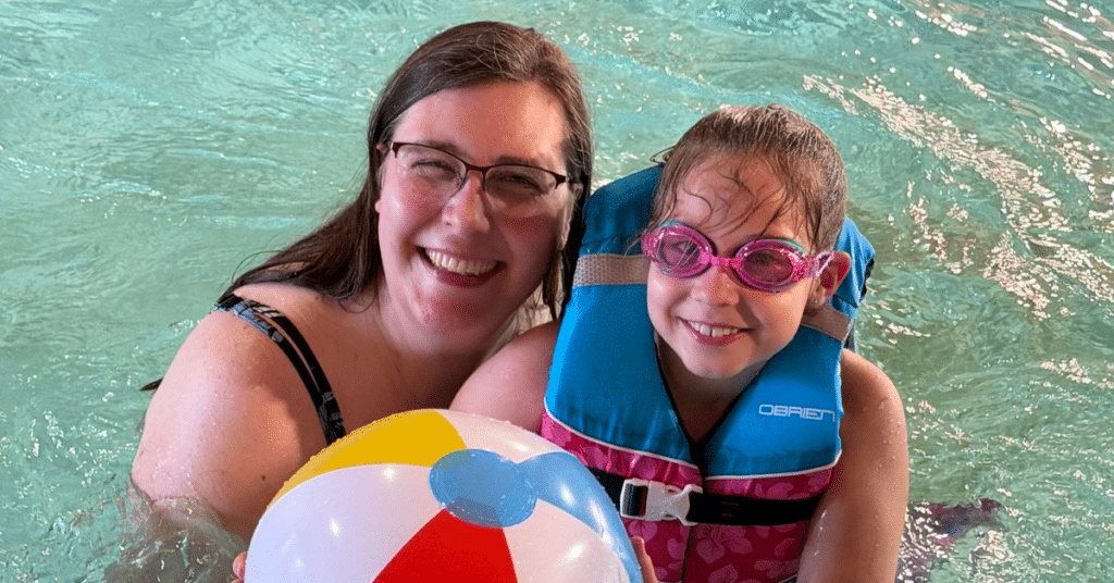 Caregiver and child pose for picture in a pool wearing a life vest, goggles, and holding a colored beach ball