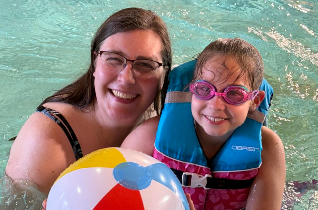 Caregiver and child pose for picture in a pool wearing a life vest, goggles, and holding a colored beach ball