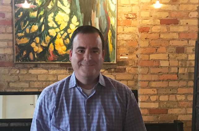 Middle-aged man looks directly at the camera in front of a brick cafe wall.