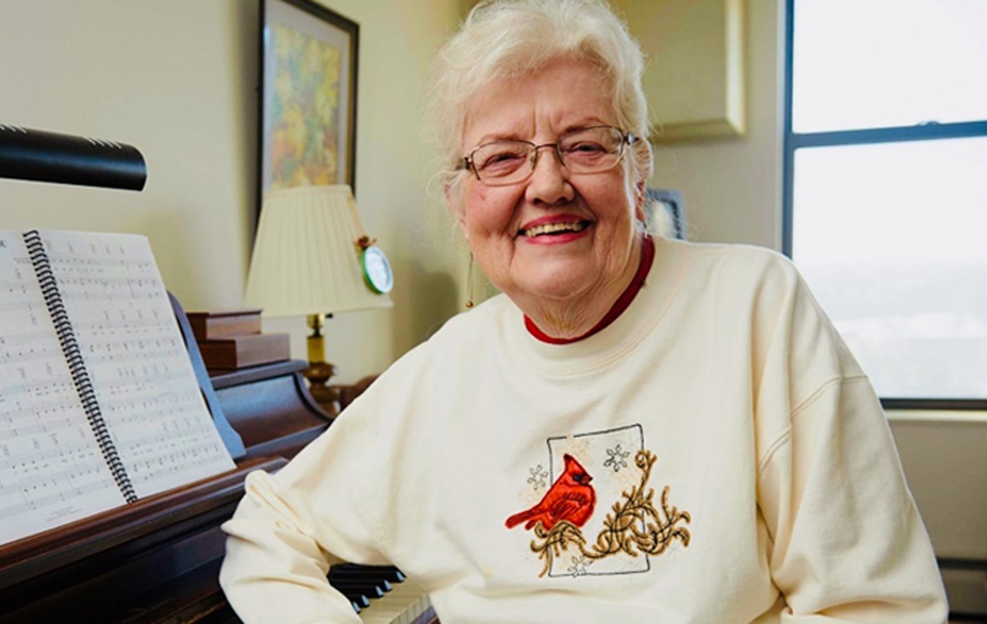 Older woman in a cardinal sweater with a big smile sits in front of a piano.
