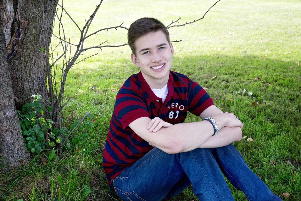 Young man sits in front of a tree, arms crossed and smiling